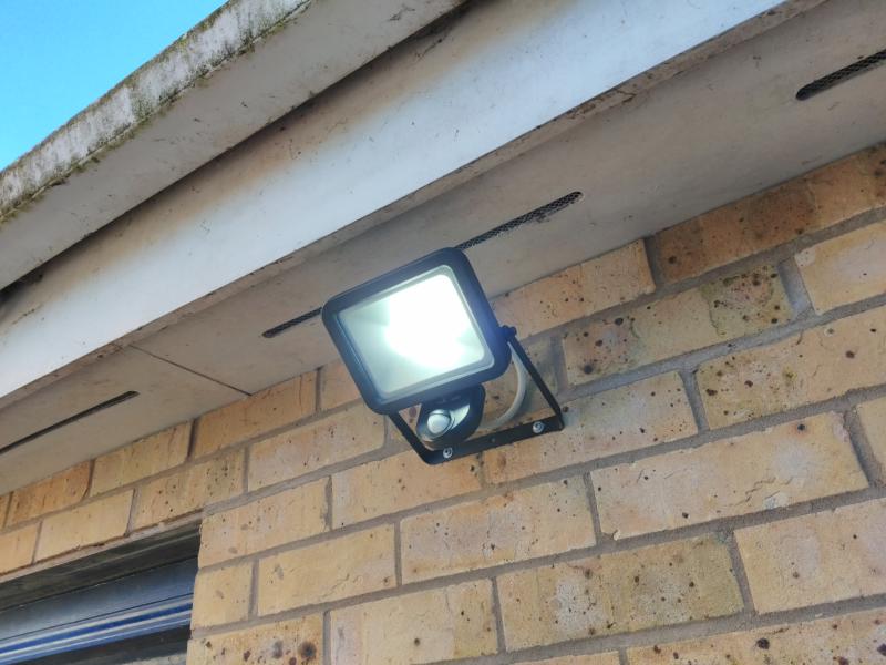 LED floodlight mounted to a brick wall, underneath some dirty/mossy gutters