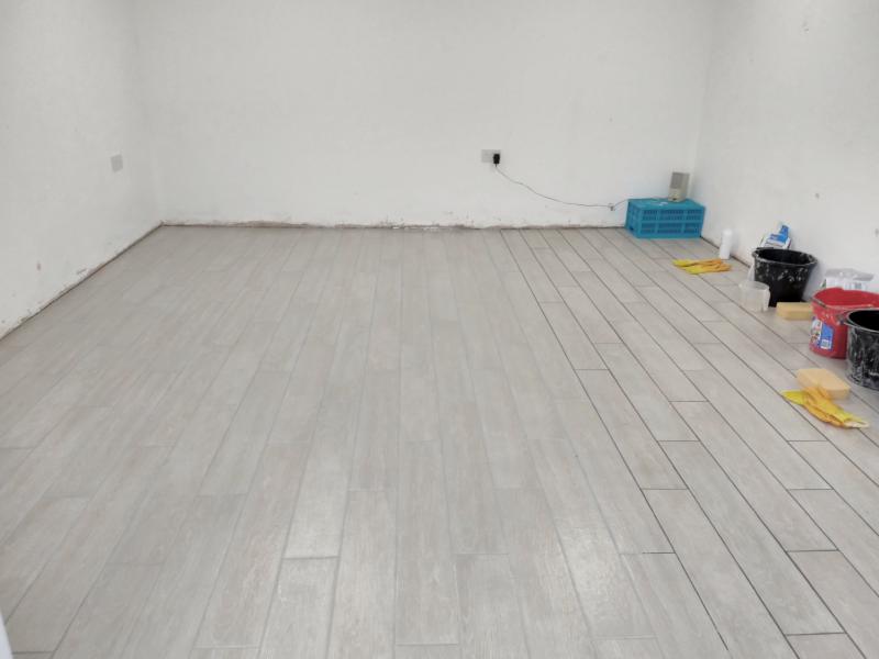 Wide view of the floor, showing about 65% of the floor having the gaps between tiles filled with a grey grout that pretty closely matches the tile colour. To the right the dark gaps are still visible. By the wall are several buckets, rubber gloves, sponges