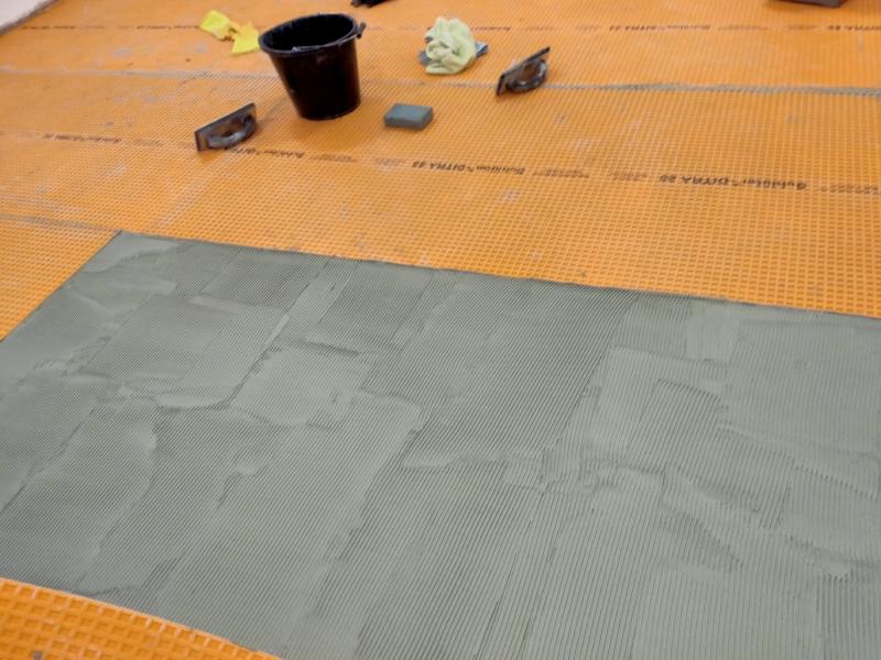 View of a section of the floor with grey mortar. The surrounding floor is covered in bright orange plastic with a square grid shape on it. The mortar has lines made of mortar ridges on it, although those sets of lines are in patches rather than continuous from one side to the other. In the background are two floats, a buckets, and some cloths