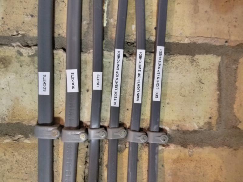 Close-up of 6 cables vertically mounted to the wall with labels on them. The leftmost 2 are thicker than the remaining 4, and are labelled 'SOCKETS'. The remaining cables are labelled, left to right: 'LIGHTS', 'OUTSIDE LIGHTS (DP SWITCHED)', 'MAIN LIGHTS (SP SWITCHED)', and 'SEC. LIGHTS (SP SWITCHED)'. A cable clip is seen on each of the cables, hammered into the same line of mortar.