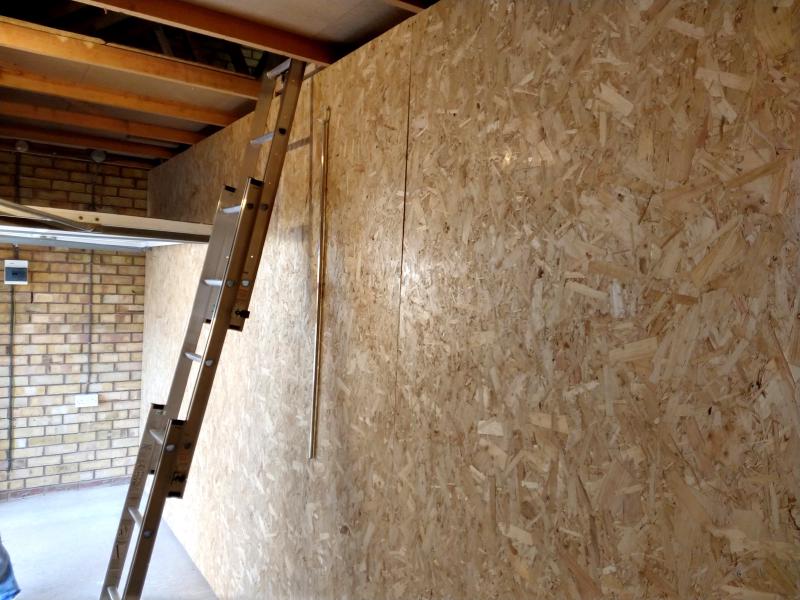 View of the front bit inside the garage; on the right is the wall dividing this front bit from the internal room. It is covered in chipboard. In the middle of the wall, a 3 section sliding ladder leans against it, leading up to the loft. Beyond, the 'up and over' garage gate is open, reaching to just a few centimetres away from this wall.