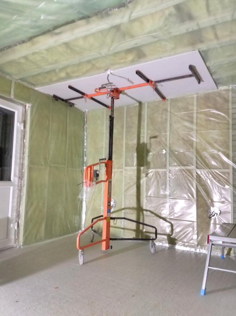 A hired board lift presses a sheet of plasterboard up against the ceiling, in a corner. The board lift is extended upwards with a large plasterboard panel on top being pushed against the ceiling. The board lift has a telescoping vertical rod which is pulled by a steel cable, operated with a rotary lever and ratchet mechanism. The part on top that holds up the board is a wide horizontal beam (also telescopic), with 4 arms perpendicular to it, evenly supporting the board