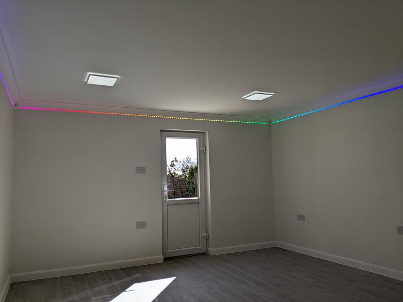 View towards the side door of the finished room. The walls are off-white, the floor grey wood-effect tile. White featureless skirting board extends around the room by the floor, and is joined to the floor and wall with white silicone sealant. The RGB LED strip underneath the coving is turned on and displays a rainbow gradient of colours. Two ceiling lights (squares emitting a white light) are visible and turned on.