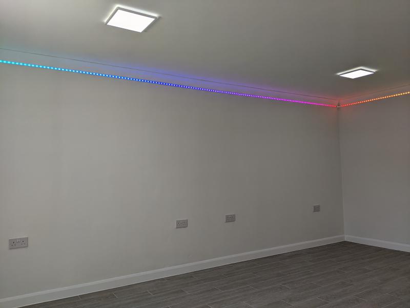 View into garage towards the same corner. The floor is made grey wood-effect long tiles, finished with plain white skirting board. The walls themselves are slightly off-white, and smooth. On the long wall toward the left, 4 double sockets about half a metre from the ground are visible. The transition from wall to ceiling is finished with white coving. Under this coving is a strip of RGB LEDs that goes all the way around, showing a rainbow gradient. The ceiling is plain white, and smooth. On the ceiling are 4 square LED light fixture which are powered on, thought only two are in the frame.
