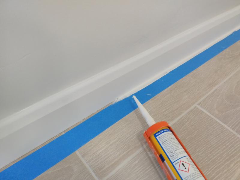 Close-up of a skirting board with blue masking tape a few millimetres away from it on the floor. On the right half, the gap between skirting board and tape is covered with smoothed over silicone sealant. In the foreground is the tip of a sealant gun pointing at it