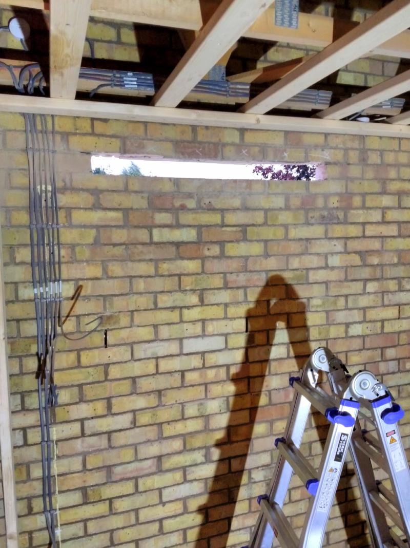 View of side wall; two rows of bricks underneath a wooden beam, 5 bricks in a row have been removed, leaving a gap through which the sky and some trees are visible. To the left of the photo are a bunch of labelled cables, though the labels are not readable from this distance. Next to the gap in the wall, the cables are covered in brown dust from the angle grinder action.