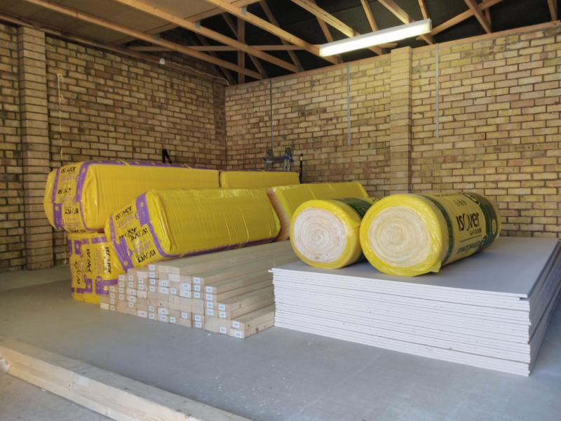 Wide view of the garage showing piles of building materials: On the right is a stack of plasterboard sheets, each 1.2 by 2.4 metres. To the left is a stack of 76 3 metre long wooden beams, each 38 by 89mm. Covering it all are packs of glass wool insulation.
