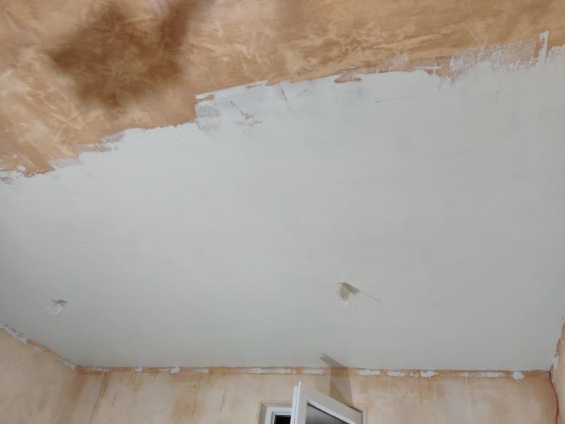 View of the ceiling, where about half of it is covered in a white coloured, very even looking plaster. Beyond the edge in the middle of the ceiling, there is uneven, patchy brown plaster instead. In the middle of the white-plastered zone are two plastic bag containing the wiring of the ceiling lights. On the walls near the ceiling are small dabs of spilled white plaster.