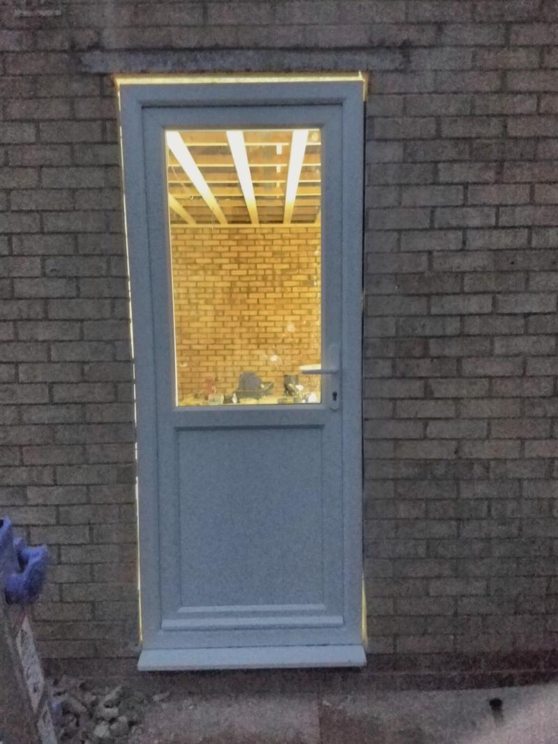Door hung in the hole in the wall. The door is a white PVC door with the top half (or slightly more) being a window. On the right is a door handle and key cylinder. The door is closed, but it's visible that it opens inwards. Around the frame of the door on the left, top and right, light from inside the room comes through gaps between brickwork and the doorframe.