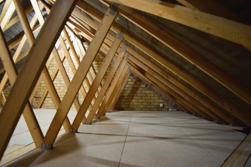 View from beyond the furthest trusses, where there is just enough height to sit. Toward the left, V shapes of the trusses , and on the floor, crisscrossing shadows. On the far wall, a junction box and power supply (for LED lighting) are seen