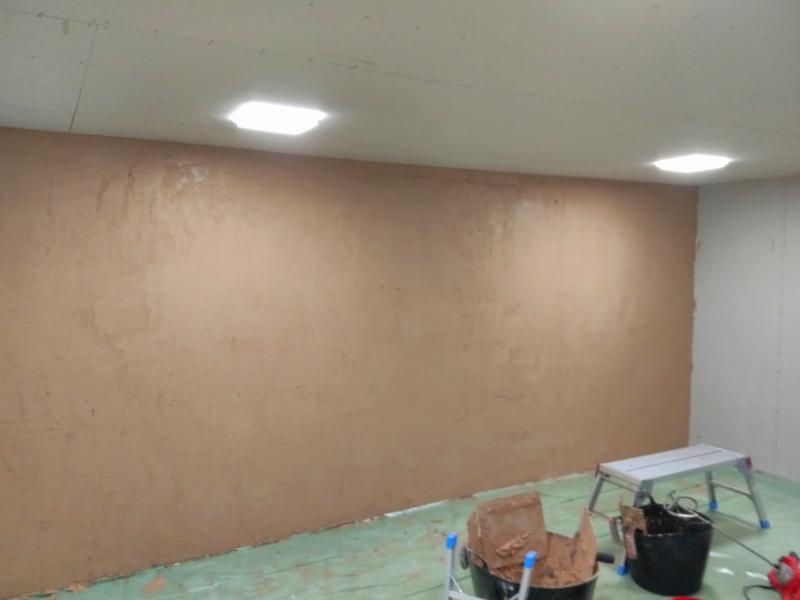 View of plastered wall, still wet. The walls is a mocha brown, in a mostly even tone. In the foreground are two mixing buckets, one of which has two hawks and a trowel perched on its edge, all covered in plaster. The other mixing bucket looks cleaner and has two more trowels perched on it, and the blade of the powered mixer leaning on it.