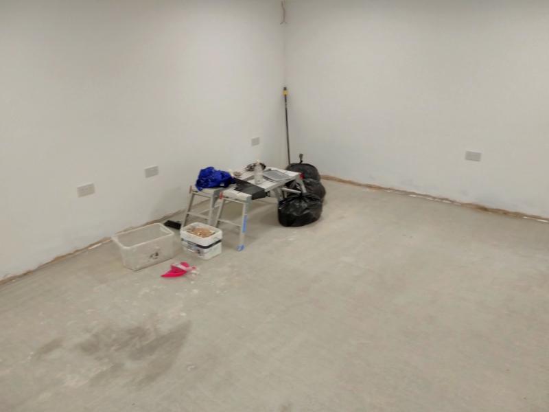 View of the floor, now without the green sheeting. The cement floor is visible, and in the corner are two cleaned work platforms, three tied rubbish bags, and a couple of rectangular contains with various waste. There's also a broom and a dustpan with brush.