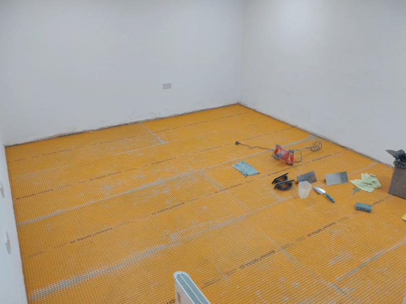 Wider view of the floor in the garage, now fully covered with the orange floor decoupling layer. Where pieces of the material meet, there is some grey mortar colouring on the edges. The floats, trowels, some cloths, a mortar mixer, and a rubbish bin and laying on the floor towards the right, on the bottom edge of the photo is an electric heater.