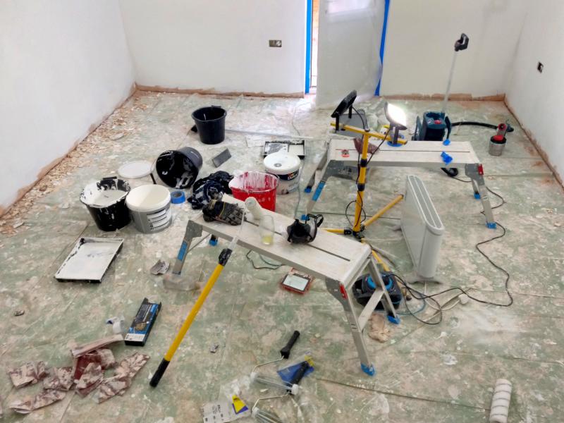 View of the floor after plastering and painting. The floor itself is covered in translucent green leftover vapour barrier sheet. This sheet is covered with stains of plaster and paint alike. Further on the floor are discarded sheets of sandpaper, Several buckets (some with paint), painters trays, a pack of paintbrushes, a single work light with some plaster covering it, a work light on a stand, two work platforms, an electric heater, and extension lead and its real, some old paint rollers and their packaging, a spray paint gun, a respirator mask, gloves, masking tape, and a paint roller telescopic pole.