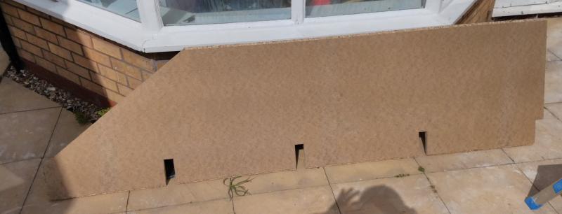 A floorboard leans against a wall. The board has its top left corner cut off at an angle of roughly 30 degrees, for a width of little less than a metre. On the bottom edge are 4 rectangular gaps, the rightmost of which extends to the right edge of the board.