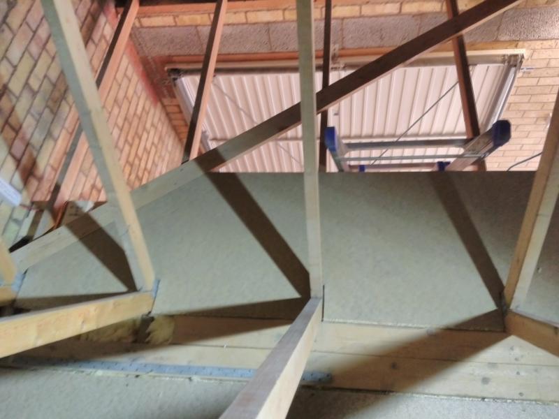The same floorboard, installed in the loft, seen from above. The diagonal cut fits snugly against a a plank that's part of the roof structure, while the gaps in the near edge fit around where trusses meet the joists the board lays on. Behind / below the floor is a closed garage gate.