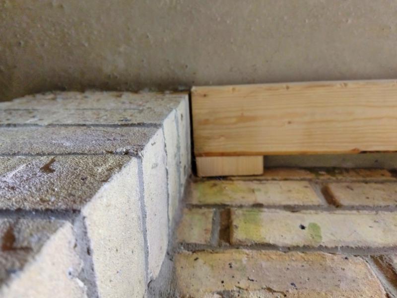Close-up looking at the floor showing a column of bricks protruding from the wall. On the floor is the end of one piece of lumber, with  small wooden off-cut holding it roughly 2-3 centimetres away from the brick wall. The wood extends about half a centimetre beyond the protruding bricks