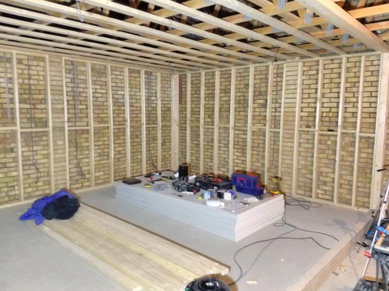 Wide view of the garage, showing wooden framework against the walls and ceiling. The framework is filled in with vertical beams further reinforced with smaller staggered horizontal beams. On the floor is a stack of plasterboard covered with various tools; next to that stack is a relatively small stack of wooden beams, 3 metres long each.