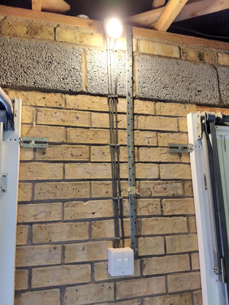 Indoor brick wall with a lit up light bulb at the top. 3 Cables lead down to a switch unit with two switches below it. The edges of the garage gates are seen on left and right edges of the photo.