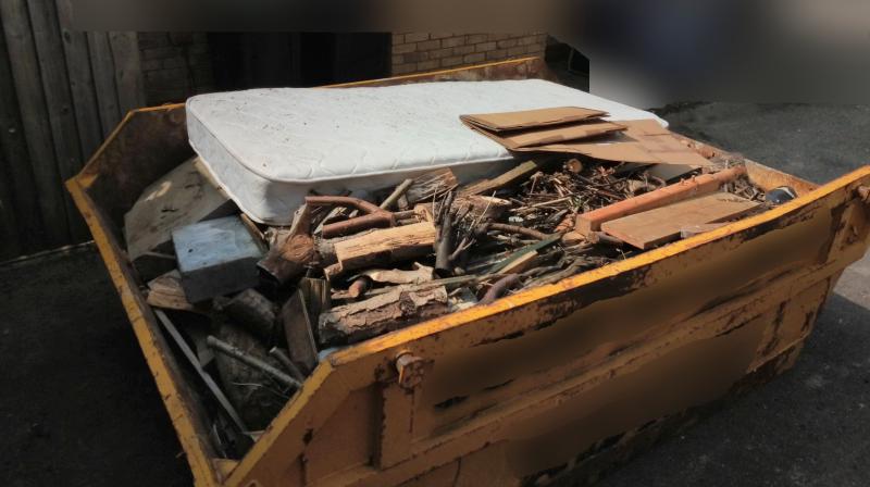 Picture of a skip, container filled with various things like a slab of stone, various bits of firewood, processed wood, cardboard boxes, an old mattress