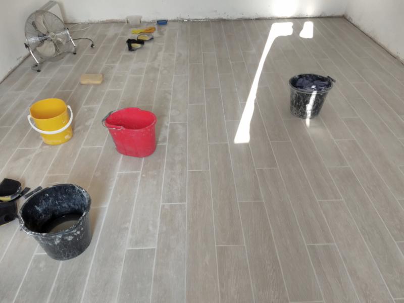 Wide view of the floor; on the right half, the tiles are clean and smooth, while on the left they are covered in grey group residue stains. Over on the dirty side are 3 buckets, knee pads, gloves, a sponge, an emulsifying pad, and a fan. On the clean side is a bucket containing used paper towels.