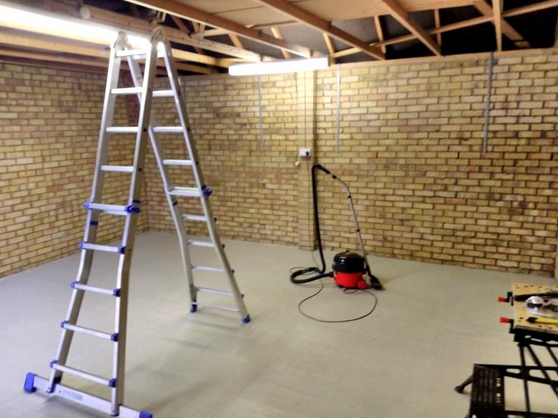 View of the new screed floor in the garage. It is now a much lighter grey and looks quite even. On the floor is a ladder in A shape under the opening in the ceiling. There's also a 'Henry the hoover' hanging out further back.