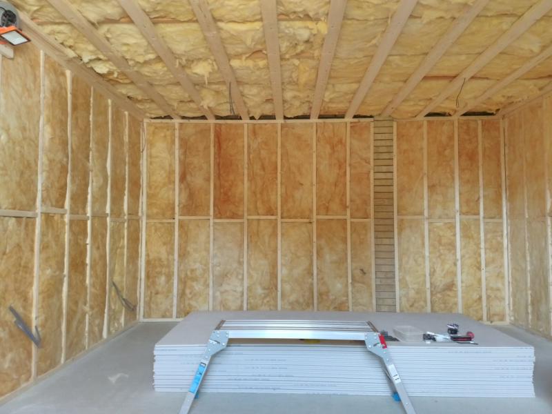 Overview of garage seen from side door; the walls are filled with insulation, and so is the ceiling. The ceiling insulation is a lighter shade of of yellow and looks less flat; lots of small pluckings protruding etc. In the top left corner, a worklight is mounted to the wooden framework. In the bottom centre on the floor is the stack of plasterboard, with only a small amount of tools left on it. In front of it stands a work platform.