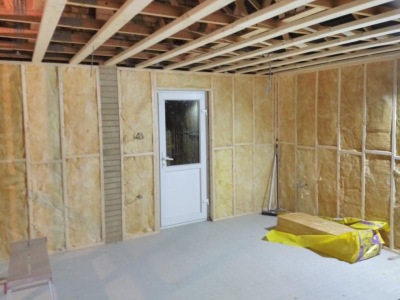 View of the left wall (with side door) and rear wall. In the wooden framework , all the holes are filled with yellow glass wool, with some electrical cable sticking through in a few places. On the floor toward the right is an opened pack of insulation with about 5 slabs left in it.