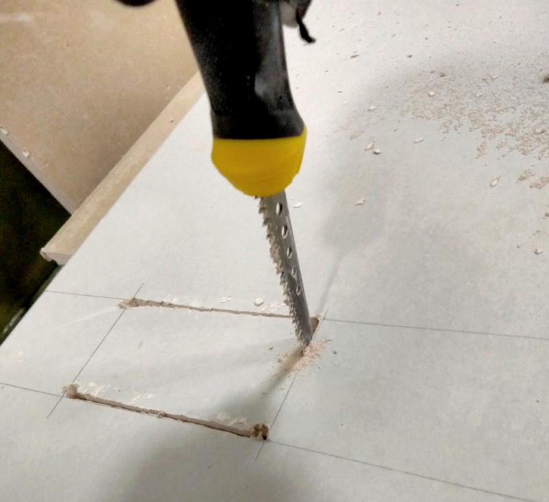 Close-up of a jab-saw cutting out the shape of a back box from plasterboard. An outline is drawn in pencil. The top and bottom cuts have already been made. The right vertical cut is underway.