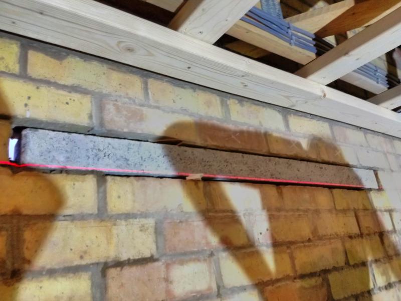 Close-up of a concrete lintel placed in the gap in the brickwork. On the bottom edge of the lintel, there is a red, laser-projected line from the laser level. Near the middle, there are some plastic shims underneath the lintel. On the left and right edges, some mortar is pushed into place.