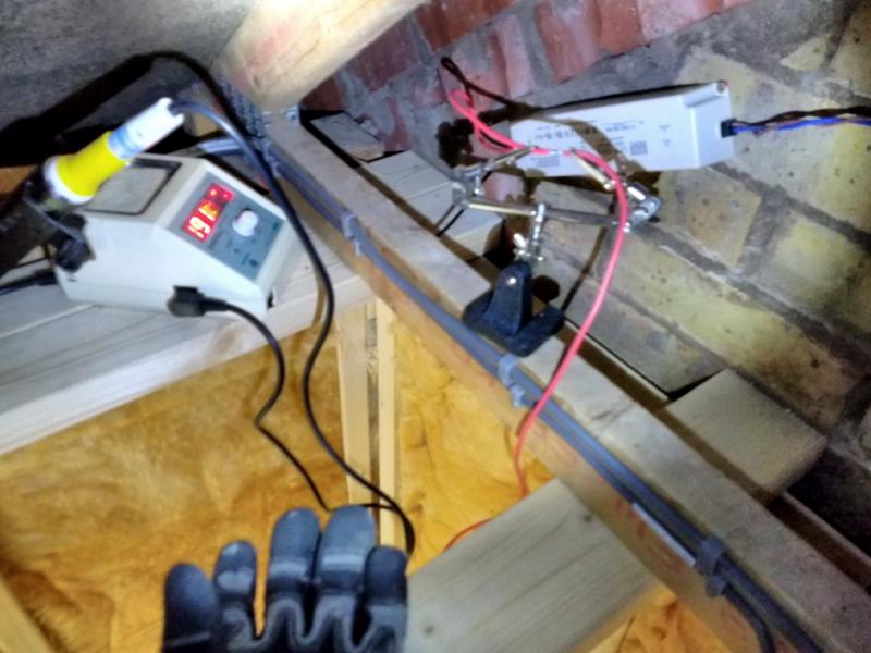 View in the very corner of the garage, above the ceiling, right under the roof. Two wooden beams side to side serve as a table for a soldering iron station. A perpendicular roof joist has a couple of cables mounted to it with cable clips. On this joist is a 'helping hand' tool: a weighted based with an adjustable pair of crocodile clips. The crocodile clips are holding two wires, so that the stripped ends are held against one another for soldering. Behind all this is a brick-shaped white plastic box (an LED power supply), screwed into the brick wall at a slightly diagonal angle. From the right end extend spiralling blue and brown wires, towards a junction box off-screen.
