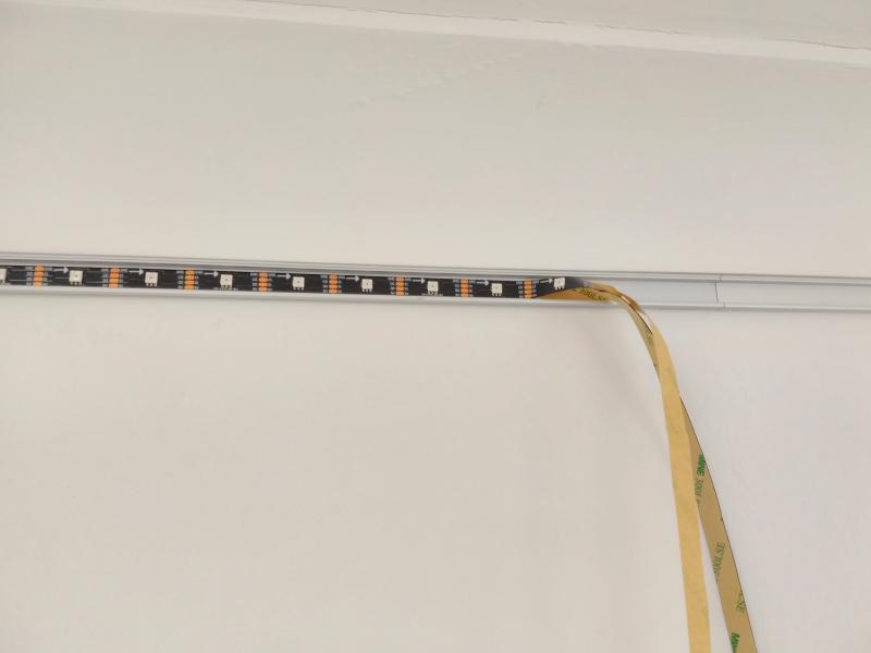 Black LED strip taped into aluminium profile. Toward the right, the LED strip with separated backing strip dangles