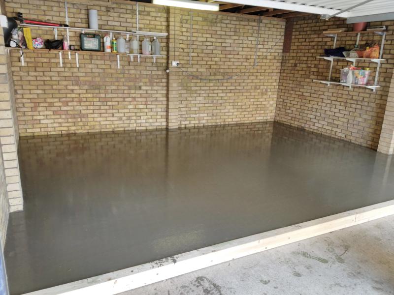 View of the area of the floor behind the wooden beams: Instead of the dark green there now is a thick layer of dark grey, still very wet screed. the wall reflects on the surface on the floor creating a depth effect.