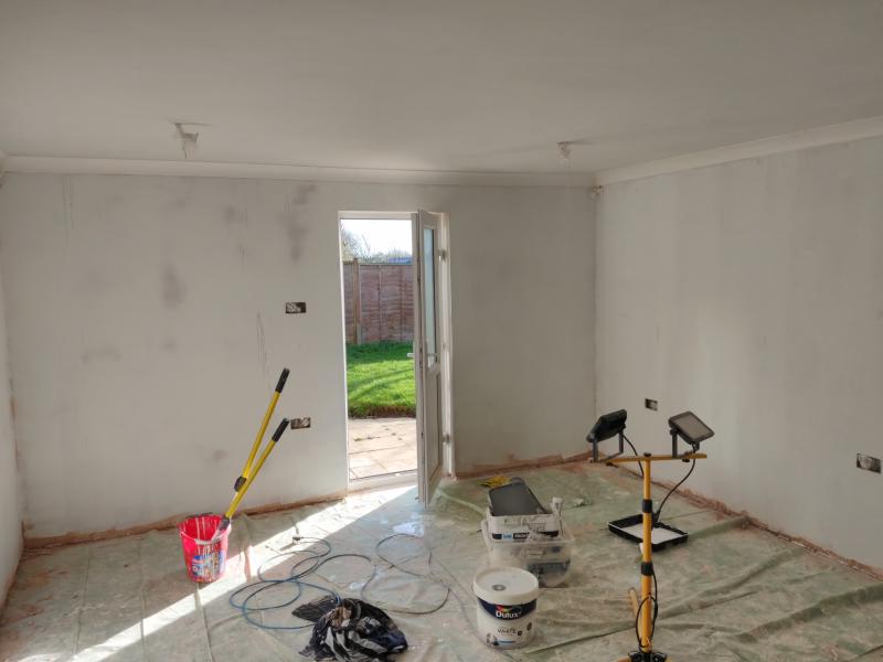 Photo of the wall with the side door, which is open. The walls are painted white with a mist coat, so it's not a solid covering. In a few places, there are darker areas in the shape of liquid spills running down the wall. In the foreground: a red bucket with two paint roller telescoping handles sticking out of it, an extension lead, a bucket of paint, a work light, a plastic box containing various waste, a paint tray, and a pile of some paint-covered clothes.