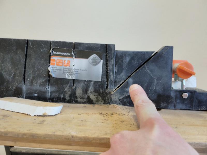 Side of a mitre box, showing 3 vertical saw blade slots, as well as one at a 45 degree angle relative to the horizontal surface. My finger points at the latter diagonal one