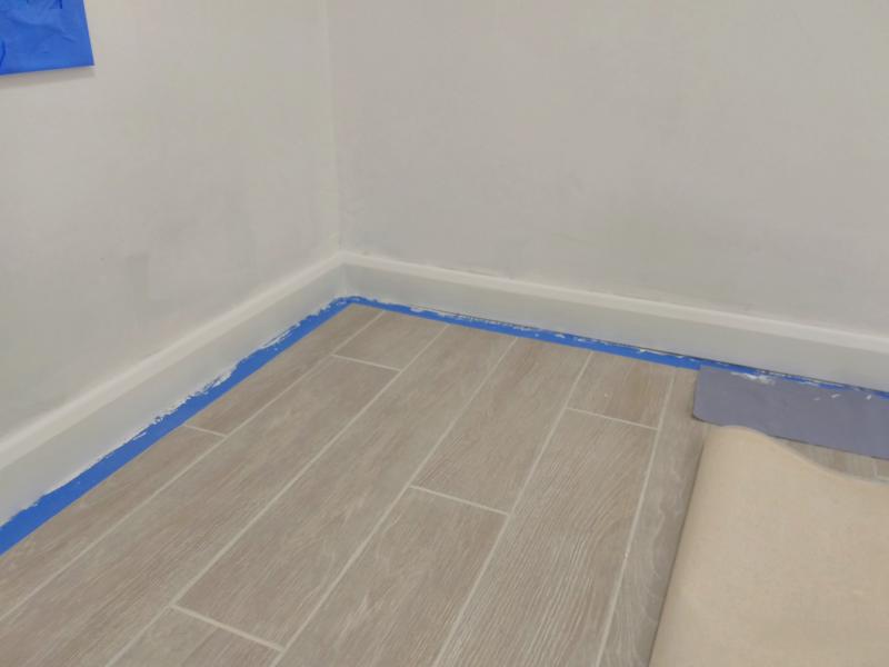 Corner of the room showing freshly painted skirting board. Next to it on the floor is blue masking tape with bits of paint all over.