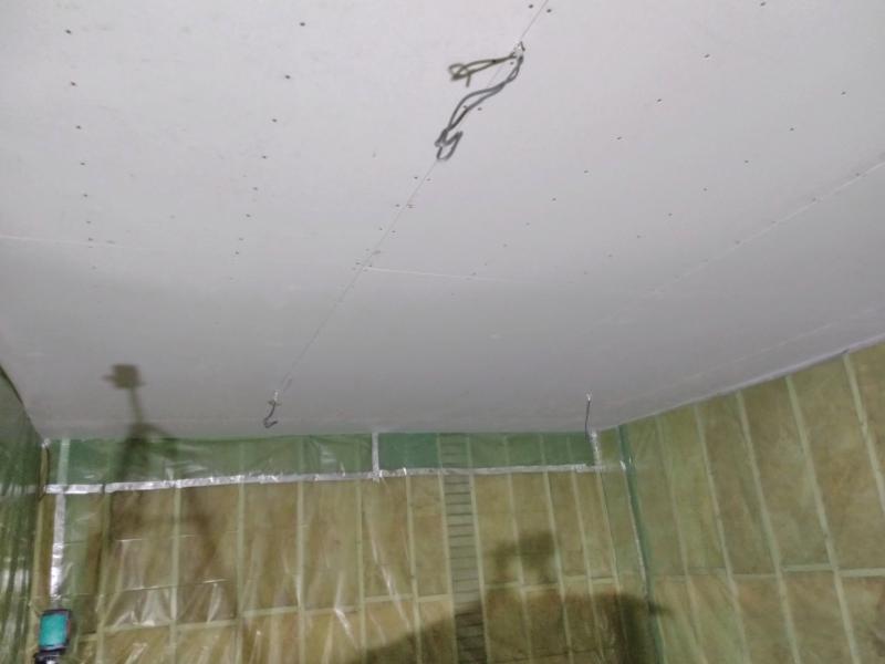 View of the ceiling now fully covered in plasterboard. Cables can be seen sticking out. The walls are still wooden framework, filled with insulation and covered with translucent green plastic foil.