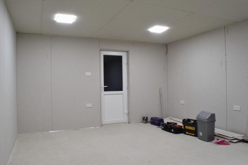 View towards the side door. Plasterboard pieces are on the inside of the doorframe, as well as above it and on the walls all around. In the top right corner of the room, a black and red wire dangle. To the left of the door is a set of 3 switches, and underneath it by about a metre is a double socket. On the right is the stack of plasterboard offcuts, and the toolboxes and bin from the last picture.