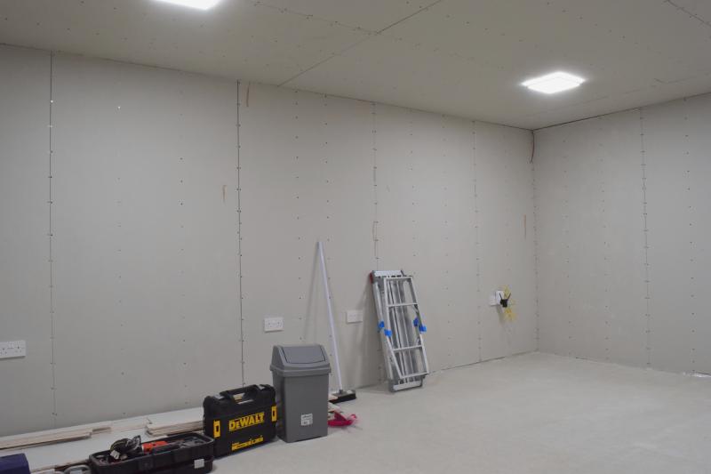View of the garage towards a corner. Plasterboard is now installed against all the walls and ceiling. Two square light fixtures are seen, both on. On the long wall there are 4 double sockets, the rightmost of which has a powerline adapter and a WiFi access point plugged into it, which are connected with a dangling yellow cable. Toward the left is a stack of plasterboard offcuts, various toolboxes, and a bin. A folded up work platform and a room are leaning against the wall.