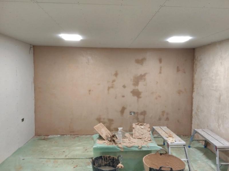 View of the second, shorter wall, plastered, still wet. The wall to the right of it is also plastered and has a much lighter shade. The newly plastered wall has a few patchy areas where the plaster looks darker than the rest of the wall. In the foreground is our coffee table as mentioned, with some plaster, two hawks and trowels, and a spray bottle on top. There are two mixing buckets and two work platforms on the floor. The floor itself is covered in leftover green vapour barrier sheet, to prevent spilling on the cement floor.
