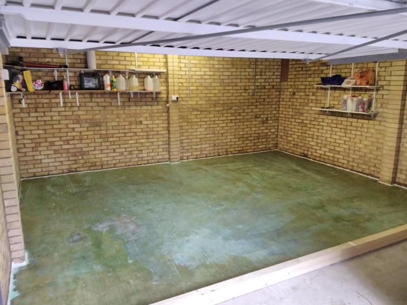 View of the area of the floor behind the newly installed wooden beams; the concrete floor has a dark green translucent covering where it's been covered with PVA. All around the edge, a white line of caulk stands out.