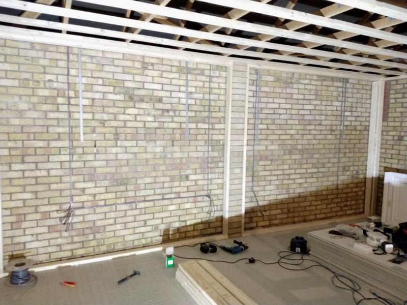 View of the rear wall, where 4 vertical cables can be seen mounted to the brickwork. On the left half, there is a horizontal cable connecting the two vertical ones as well. On the floor is a roll of cable, a pair of side cutters, a hammer, a small cardboard box, hearing protection headphones, a mains powered drill.