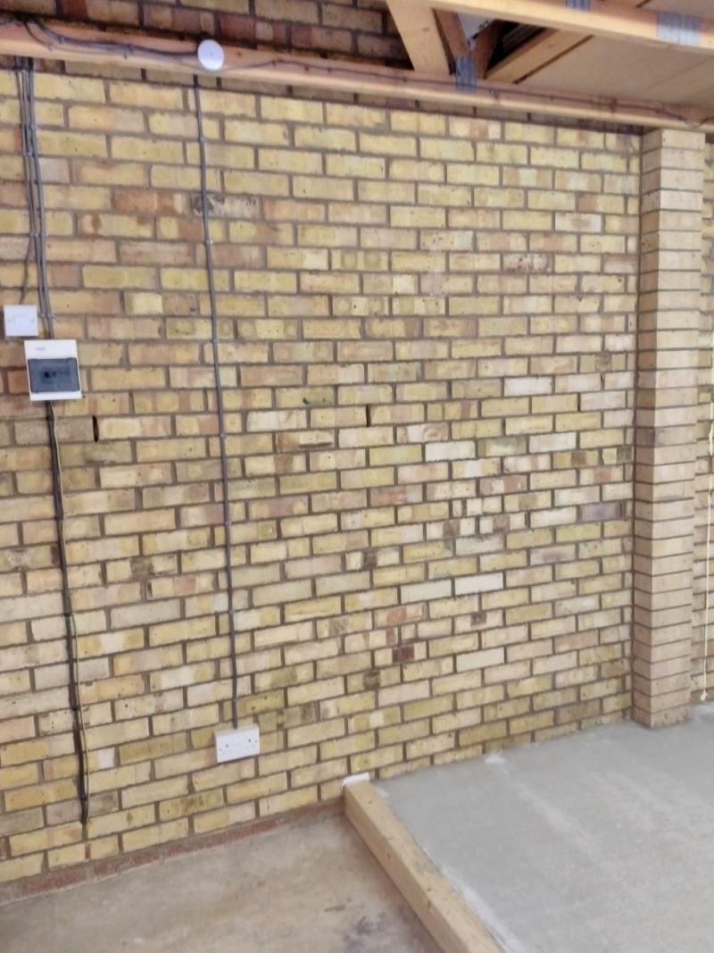 View of the left brick wall, showing a newly placed double socket near the floor. A cable leads up from it, to a junction box mounted on a wooden beam. To the left is a consumer unit (breaker box) serving the garage