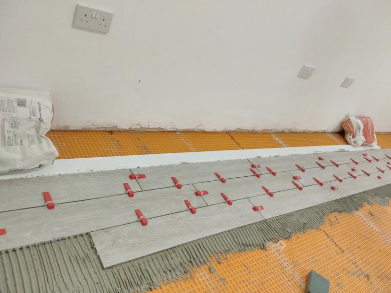 A few rows of long rectangular tiles are laid out on mortar with ridges in it. The tiles are lined up against a white piece of skirting board held down with two bags of plaster. Between the tiles are translucent spacers, each with a red wedge pushed through over the top of the tiles.