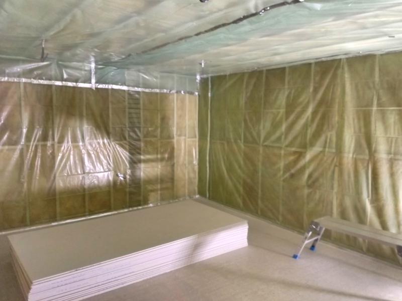 Wide view of garage, showing walls and ceiling fully covered in translucent green plastic sheeting, and the joints covered in reflective tape. The cement floor is clear safe for a work platform towards the right, and a stack of plasterboard.
