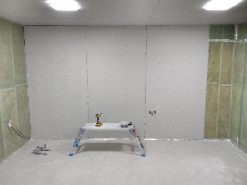View of the right wall of the garage. 3 Plasterboard panels have been installed, starting from the left edge. On the right end of the wall, there is still about half a metre of uncovered insulated wall. In from of the wall there's a work platform with an impact driver, a bucket of screws, and a laser distance measuring device. On the floor to the left there are two metal board lifts. The board lifts are a horizontal plate of metal with ridge near the end (to push against the plasterboard.) Underneath this metal plate is a ridge it balances on, used as a pivot point for the lever action. Over the top is a loop to fit around the user's shoe, to easily carry it around hands-free.