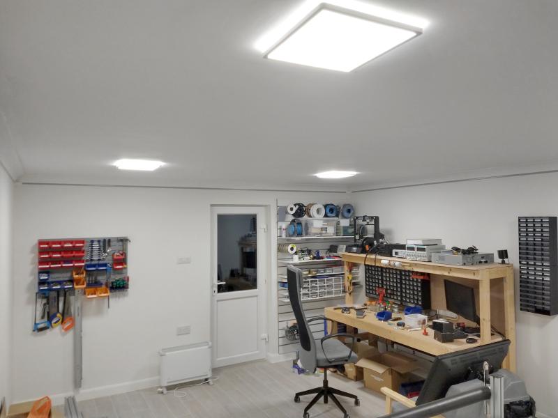 Wide view of the room. In the right half is a wooden workbench with a shelf on top. The shelf has various electronics test equipment and a 3D printer. On the bench there is a computer, several storage bin kits. To the left of the workbench is shelving made from slatted wall. To the right of the bench are more part bins mounted to the wall. To the left of the slatted wall is a closed door with a window, and left of that an organiser mounted to the wall, sporting a variety of hand tool, drill bits etc, and a bunch of bins with screws etc. Hanging from it are several saws and a long ruler.