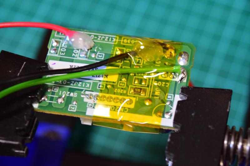 A/C circuit board clamped into PCB vise, green side up, connector towards us. In the top left, a red wire is soldered to one of the LEDs pins, and glued down with hot glue. To the top right, a black wire is soldered and glue down the same. On the right edge, a gree wire is soldered but not yet glued. It is is held onto the board with yellow clear kapton tape for soldering.