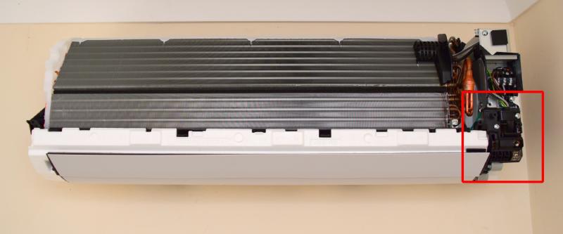Bare A/C unit on the wall. The heat exchanger, a large stack of thin metal fins takes up the bulk of the size. On the right are some copper pipes, screw terminals and wiring, and in the bottom right (marked), a plastic assembly containing the infrared sensor and other electronics