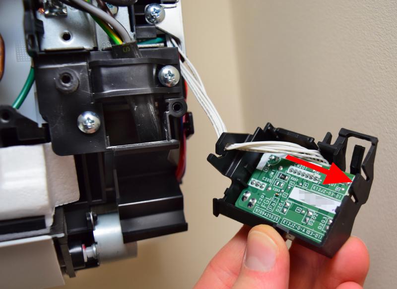 The module removed and turned over. There is a circuit board held into the plastic, with another latch on the right. A 70wire cableplugs into the obscured side of the PCB. Its wires are routed through a hook in the left of the plastic assembly and lead off into the A/C unit. An arrow indicates the plastic latch on the right, and the direction to push it to remove the board.